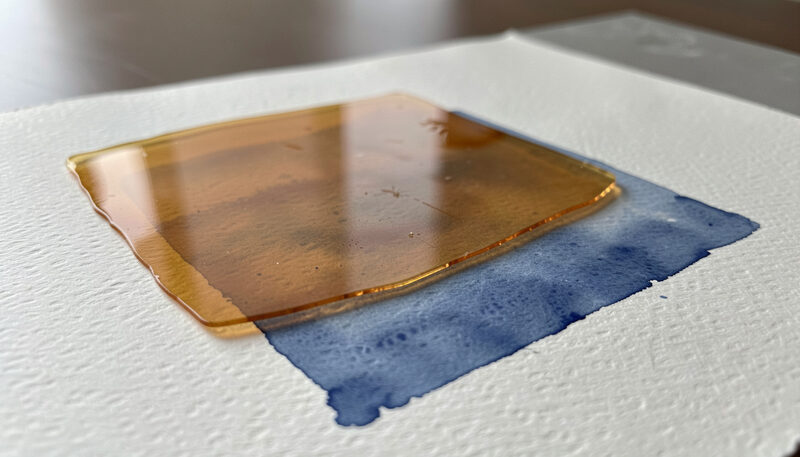 Close view of thin transparent layers of warm ochre drying over a blue underpainting on textured paper.