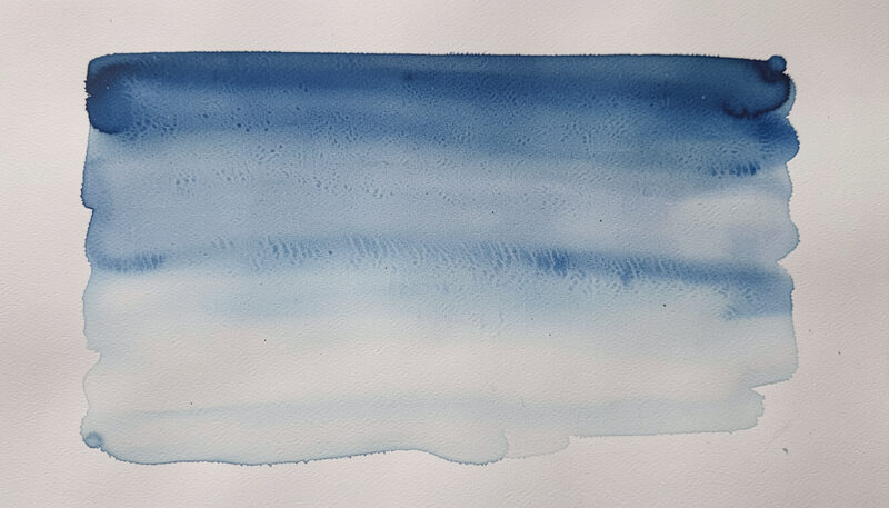 A shallow-tilted wooden drawing board on a plain desk, taped watercolor paper with a single graded wash drying, a loaded round brush resting on a ceramic palette beside two glass water jars, side lamp casting a soft shadow, close three-quarter view in daylight.
